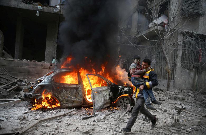 Thủ đô Damascus của Syria bị tàn phá nặng nề trong các cuộc giao tranh tiếp diễn giữa lực lượng chính phủ Syria và các nhóm phiến quân. Nhiều nhà cửa đã bị phá hủy và cuộc sống người dân địa phương bị ảnh hưởng nghiêm trọng. Ảnh: Một thành viên của lực lượng Phòng vệ Dân sự Syria bế em nhỏ bị thương đi qua chiếc ô tô đang bốc cháy tại thị trấn Harmoria, Đông Ghouta, ngày 6/1. (Nguồn ảnh: Reuters).
