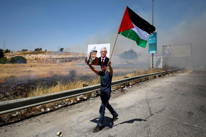 Một thanh niên cầm cờ Palestine và bức ảnh cố lãnh đạo Palestine Yasser Arafat và Tổng thống Mahmoud Abbas đi bộ trên đường phố mù mịt hơi cay gần khu định cư Beit El ngày 24/6. Ảnh: Reuters.