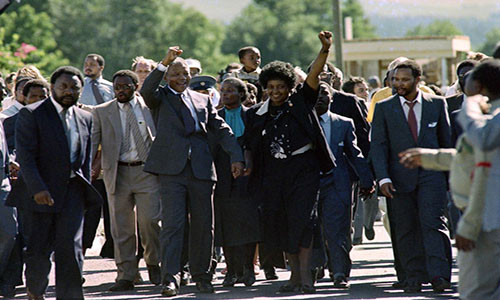 Vợ chồng ông Mandela nắm tay nhau rời khỏi nhà tù Victor Verster và vẫy chào đám đông vào năm 1990 ở Paaarl, Nam Phi. Ảnh: Getty.