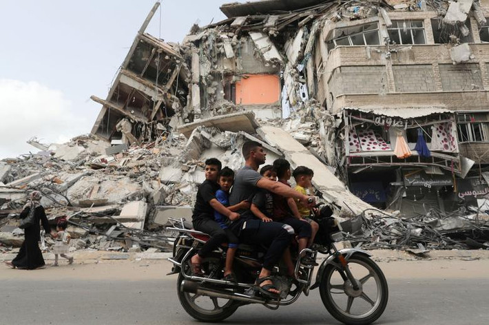 Người đàn ông chở các em nhỏ trên xe máy đi qua tòa nhà đổ nát tại Gaza hôm 21/5.