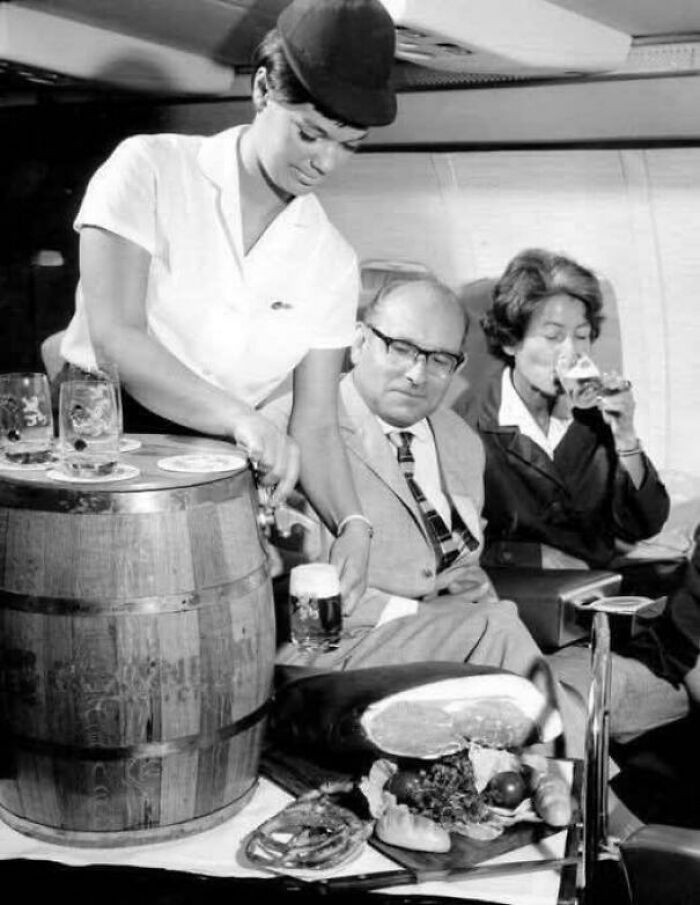Tiếp viên hàng không của hãng hàng không Đức Lufthansa phục vụ bia tươi từ thùng gỗ và giăm bông cho hành khách trên chuyến bay, khoảng năm 1963.