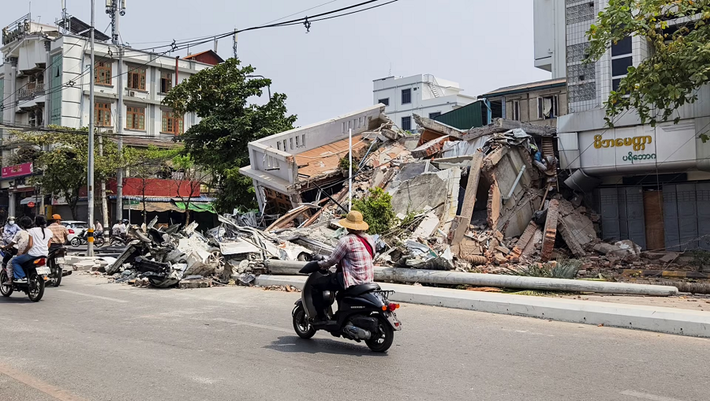 Người dân đi xe máy đi qua đống đổ nát ở Mandalay.