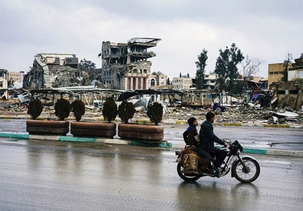 Hai người đàn ông đi xe máy qua những tòa nhà đổ nát trong chiến tranh ở Mosul.
