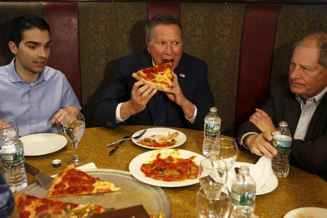 Thống đốc bang Ohio John Kasich - ứng viên của Đảng Cộng hòa không ngần ngại hình tượng, há miệng cắn một miếng pizza lớn trong khi ngồi cùng cựu Nghị sĩ Bob Turner (phải) và một thành viên hội đồng thành phố New York.
