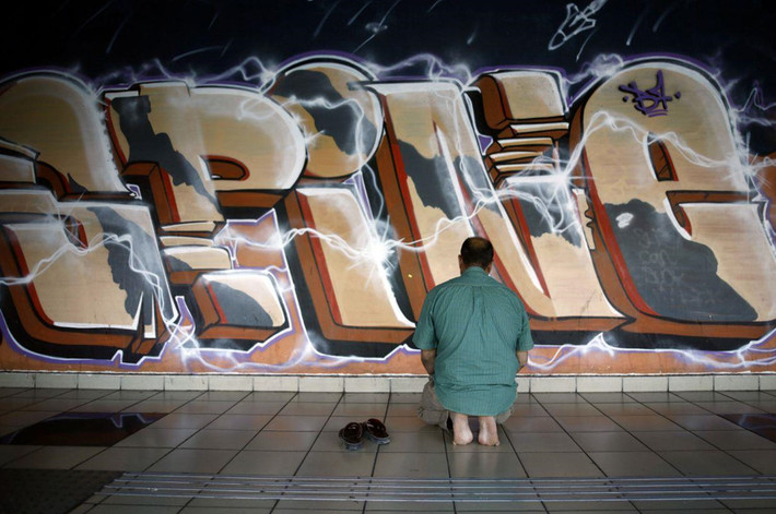 Một người đàn ông Hồi giáo cầu nguyện gần bức graffiti tại trạm xe buýt. Sự đa dạng của nhà ga phản ánh khu vực, nơi có lượng lớn người di cư nước ngoài sống trong các khu chung cư cũ.