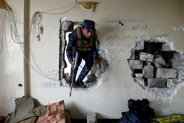 Một cảnh sát Iraq đi qua lỗ hổng trên tường để vào một ngôi nhà ở Tây Mosul.