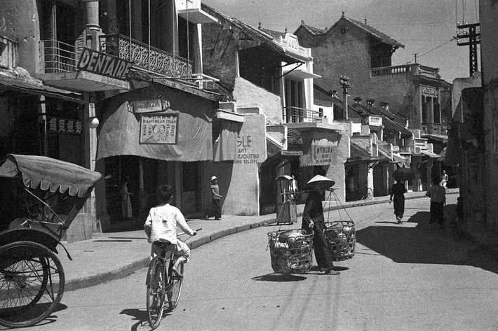 Một phụ nữ với đôi quang gánh hàng trĩu nặng trên vai di chuyển trên phố Hàng Bạc.