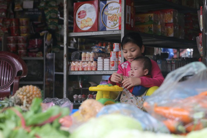 Tại một cửa hàng bách hóa trong thôn, bé gái thay mẹ vừa trông hàng vừa dỗ em để mẹ chuẩn bị bữa cơm trưa.