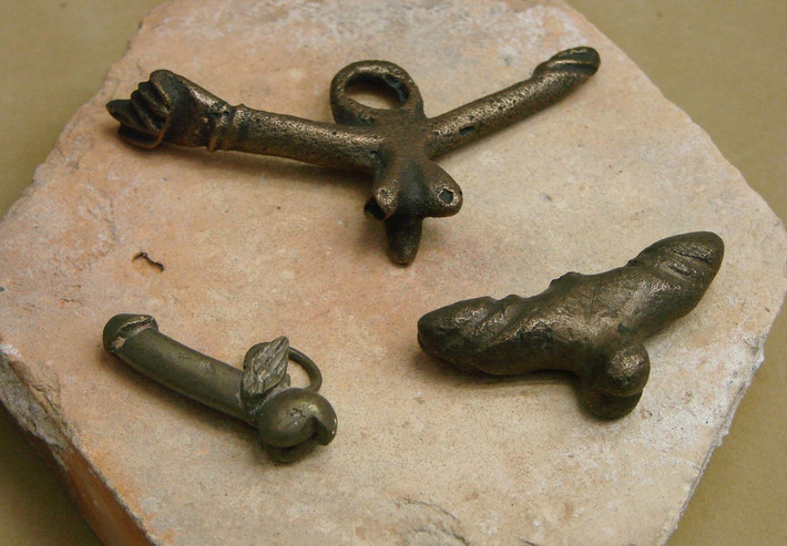 Nguyên do là bởi Marcus Terentius Varro (sống từ năm 116 trước Công nguyên - 27 trước Công nguyên) và Pliny the Elder (sống từ năm 23 sau Công nguyên - 79 sau Công nguyên) đã có những ghi chép về việc người La Mã cổ đại từng sử dụng các món đồ có hình "của quý" giống như bùa hộ mệnh để bảo vệ bản thân khỏi những điều xui xẻo, hắc ám.