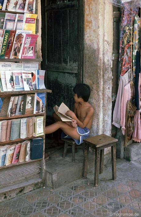 Một bé trai ngồi đọc một cuốn sách khá dày khi trông cửa hàng bán, cho thuê sách, truyện, tạp chí... cũ.