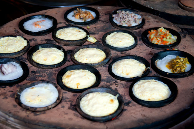 Bánh căn: Bánh căn gần giống bánh khọt, nhưng làm bằng bột gạo pha nếp, trên lò than hồng là khuôn bánh bằng đất nung có đến cả chục khuôn nhỏ giống như khuôn bánh khọt. Người đổ bánh có kinh nghiệm làm sao bánh khi ra khỏi khuôn phải vừa giòn, vừa xốp lại vừa dẻo, vỏ bánh phải có màu trắng vàng nhưng không bị cháy khét. Bánh căn được nướng chín bằng khuôn đất nung nên ăn cũng không mau ngán. Ảnh: Dulich24.