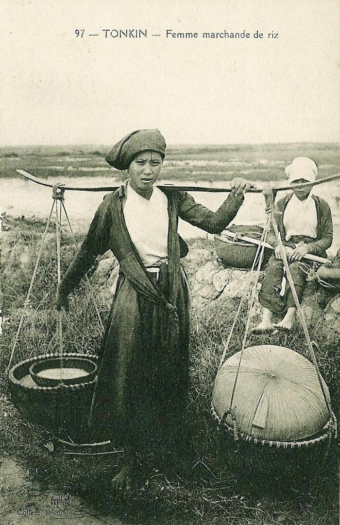 Người phụ nữ với đôi quang gánh đựng các thúng gạo trên vai khi đi ra chợ. Ở phía sau là một người phụ nữ tạm dừng chân nghỉ ngơi ven đường.