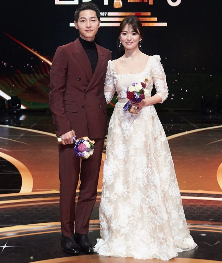 Khi xuất hiện ở sự kiện, Song Hye Kyo diện đầm trắng cổ trễ, thiết kế ren nhiều lớp hết mực thanh thoát, nhẹ nhàng. Trong khi đó, ông xã của cô chọn bộ suits màu đỏ burgundy, phối cùng áo len cổ lọ ấm áp.