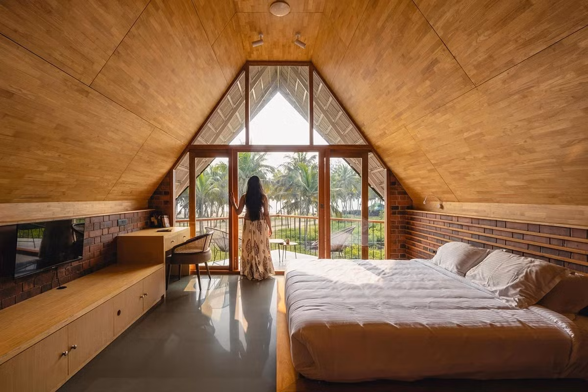 Phòng ngủ bố trí dưới mái dốc lớn, sử dụng gỗ sáng kết hợp gạch nung đỏ. Ban công mở trọn tầm nhìn hướng biển. Ảnh: Bhoomija
