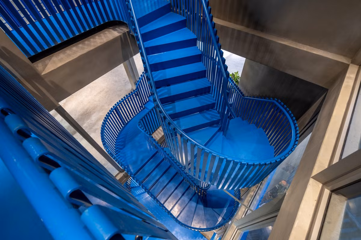 Một cầu thang xoắn ốc màu xanh lam sống động đóng vai trò là điểm neo điêu khắc của không gian, dẫn hướng chuyển động qua các chức năng phân lớp của quán cà phê, triển lãm và khu vực làm việc chung. Ảnh: Anh Chương, Bánh shimano