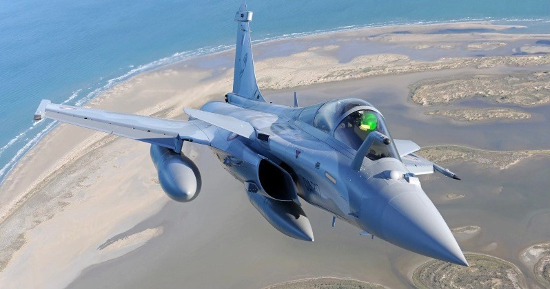 Tiêm kích Dassault Rafale bay thấp trên khu vực ven biển, phi công đội mũ bay với kính hiển thị gắn trên mũ (HMD), mang theo thùng nhiên liệu phụ dưới cánh trong một chuyến bay huấn luyện hoặc tuần tra.