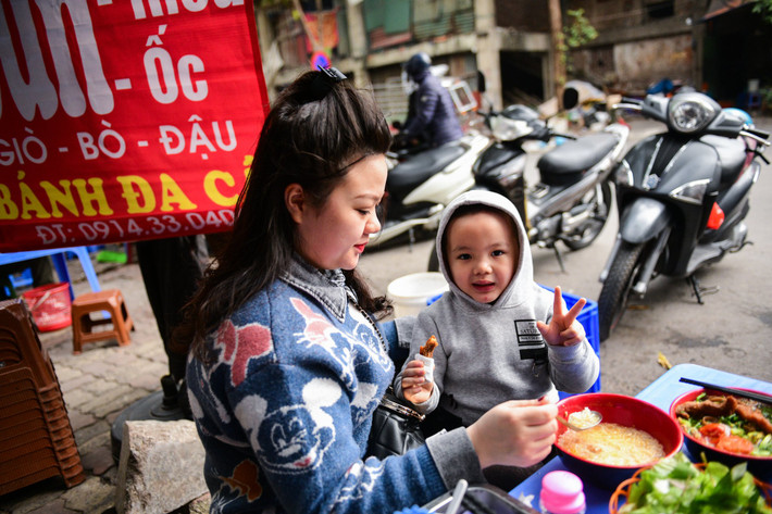 Chị Khánh Chung cùng con trai đi ăn trưa tại một quán bánh đa cá ở Đội Cấn (Ba Đình). "Mấy ngày Tết ăn cỗ ở nhà nhiều quá, giò chả, xôi, gà... ngấy lắm rồi nên gia đình tôi đi đổi bữa", chị nói. Đây là quán quen chị thường xuyên ăn, hôm nay tăng thêm 15.000-20.000 đồng mỗi bát.