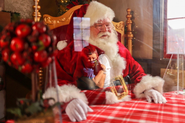 Đằng sau lớp khẩu trang, kính chống giọt bắn và vách ngăn nhựa, các ông già Noel vẫn đang tiếp tục gặp mặt các em nhỏ. Mọi năm, lũ trẻ sẽ ngồi lên lòng, nói chuyện với ông hoặc đứng sát gần. Năm nay, điều này là không thể vì cần giữ khoảng cách tối thiểu. Trong ảnh, ông Panchito Vicente (74 tuổi) đang ngồi sau tấm ngăn nhựa tại trung tâm thương mại Citadel Outlet ở California (Mỹ).
