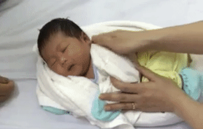 Bước 2: Em bé cần mặc áo quần gọn gàng, thoải mái. Sau đó để em bé nằm nghiêng bên phải và vòng khăn từ dưới chân lên đầu. Một đầu khăn đặt ở dưới cổ và đầu còn lại đặt trên đầu em bé.