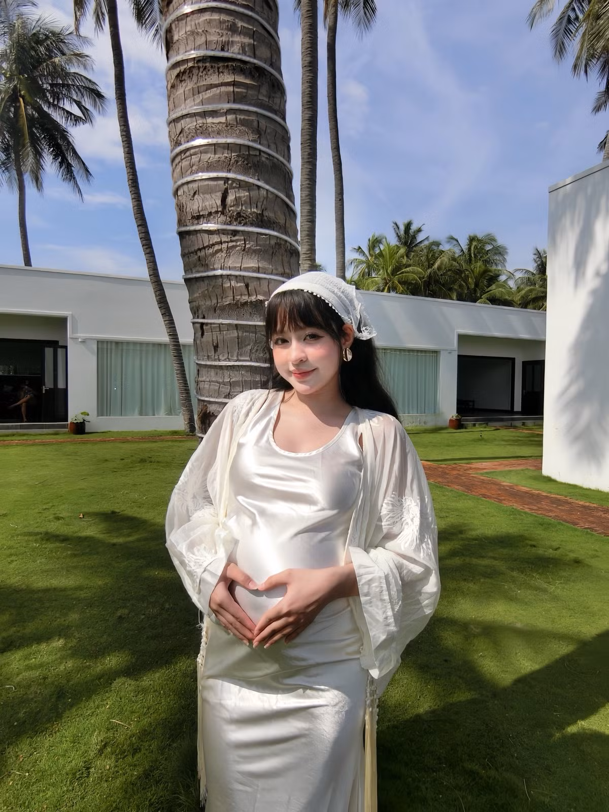 Bộ ảnh được thực hiện trong không gian ngoài trời thoáng đãng, với bối cảnh là bãi cỏ xanh mướt, những cây dừa cao vút và kiến trúc nhà nghỉ dưỡng màu trắng tối giản, càng làm nổi bật vẻ đẹp thuần khiết, tươi tắn của mẹ bầu. (Ảnh: FBNV)