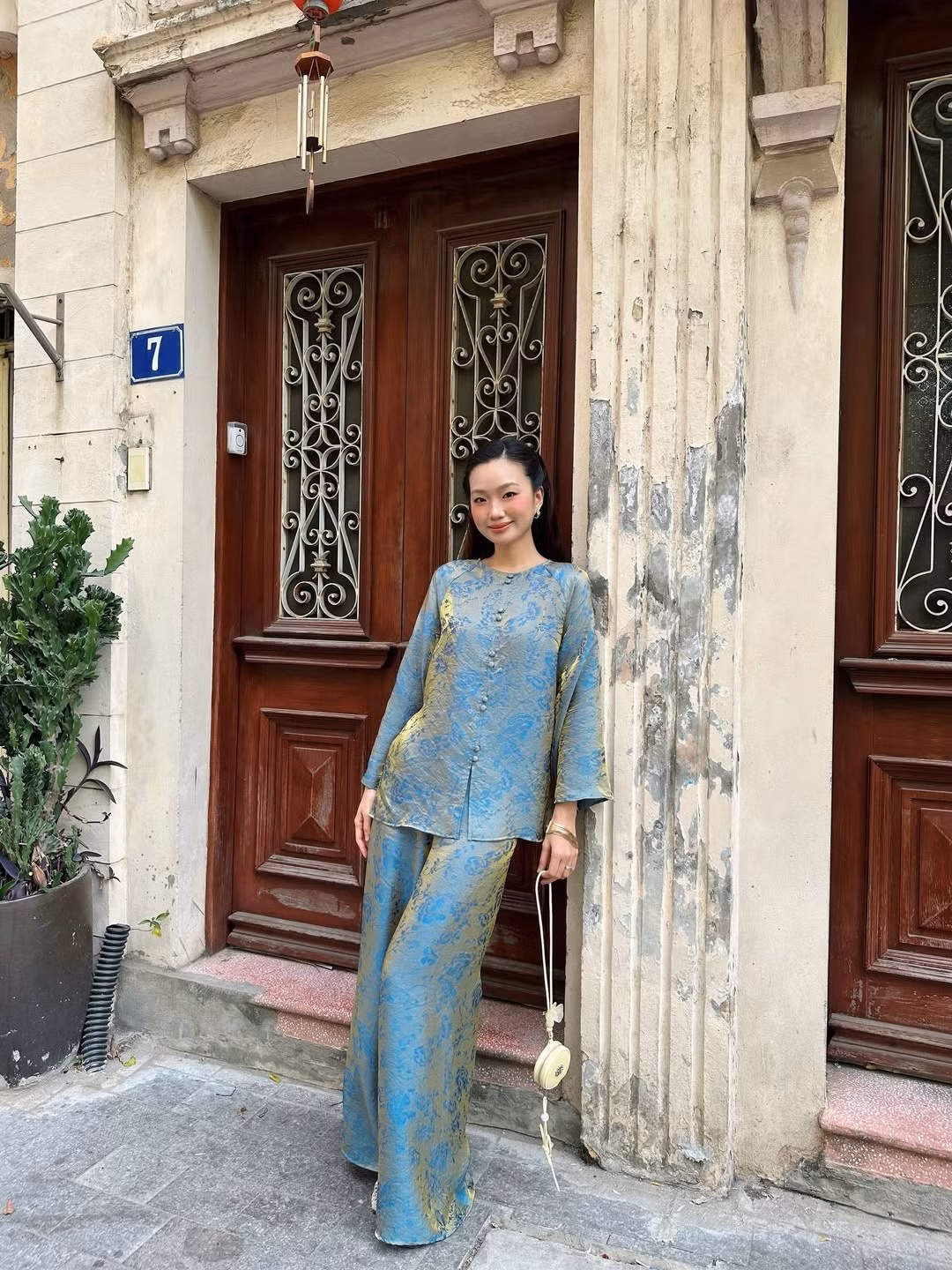 Bộ trang phục của Chái Chanh sử dụng chất liệu gấm cao cấp với họa tiết hoa chìm tinh tế. Ánh kim vàng trên nền vải xanh khói tạo nên hiệu ứng thị giác cực kỳ sang trọng dưới ánh nắng dịu dàng những ngày cuối năm. (Ảnh: IGNV)