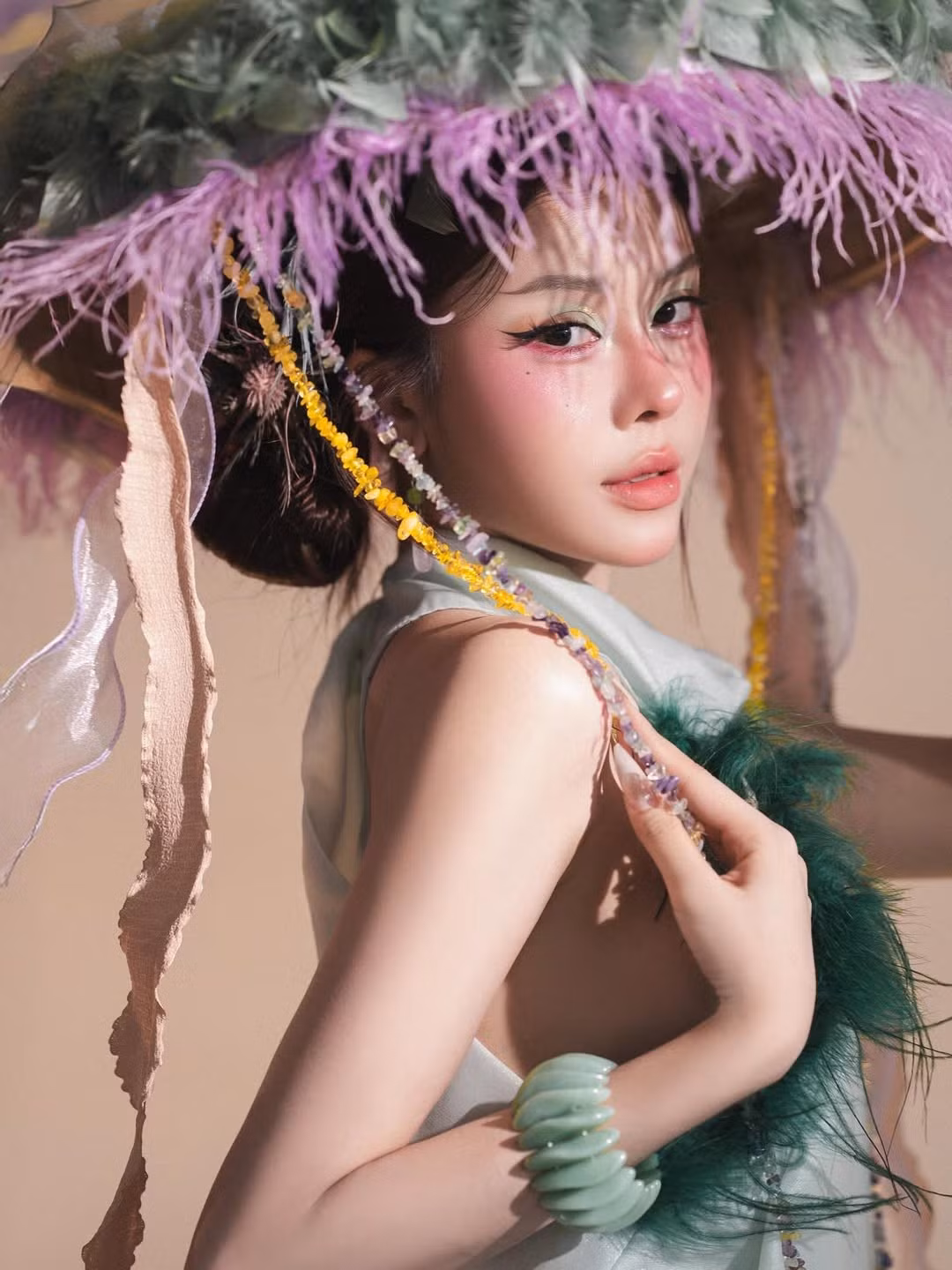 Nổi bật nhất phải kể đến chiếc nón lá cách điệu khổng lồ được đính kết lông vũ xanh lục và tím oải hương cùng những chuỗi hạt đá quý rủ xuống, làm tăng thêm vẻ ma mị, thoát tục cho tổng thể. (Ảnh: IGNV)