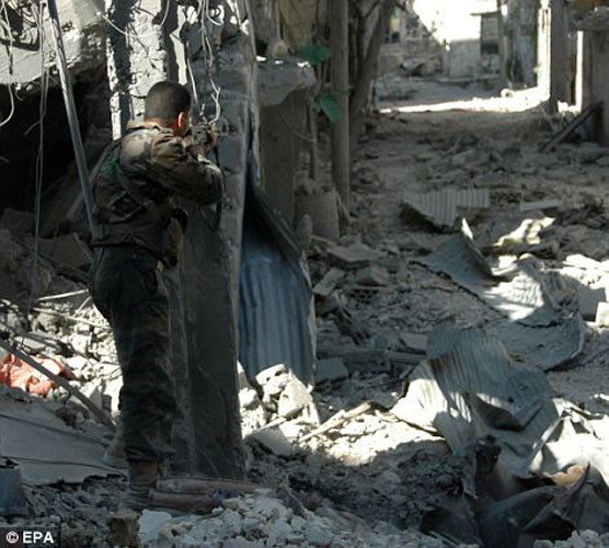 Một người lính quân đội chính phủ Assad đứng nấp đằng sau một khối bê tông xung quanh là đống đổ nát, hoang tàn.