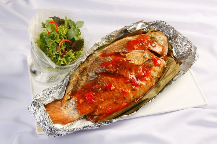 Ướp sốt trên với cá trong 1giờ, xoa đều cho cá ngấm gia vị, bọc màng thực phẩm, để tủ mát. Cá bọc kín giấy bạc, đem nướng lò 25 phút ở 200 độ C. Hành lá, ớt sừng thái chỉ, chanh thái lát mỏng, thì là đã rửa sạch. Rắc lên trên cá sau khi nướng. Ảnh: nhipsongso.edu.vn.