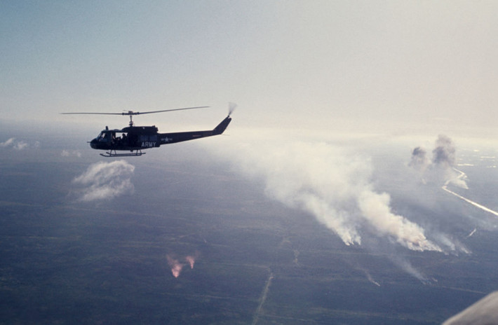 Một chiếc trực thăng UH-1 của Mỹ bay qua vùng nông thôn miền Nam thời chiến tranh Việt Nam, năm 1965. Dưới mặt đất, khói bốc lên từ những khu vực bị quân Mỹ oanh tạc. Ảnh: Jean-Claude Sauer.