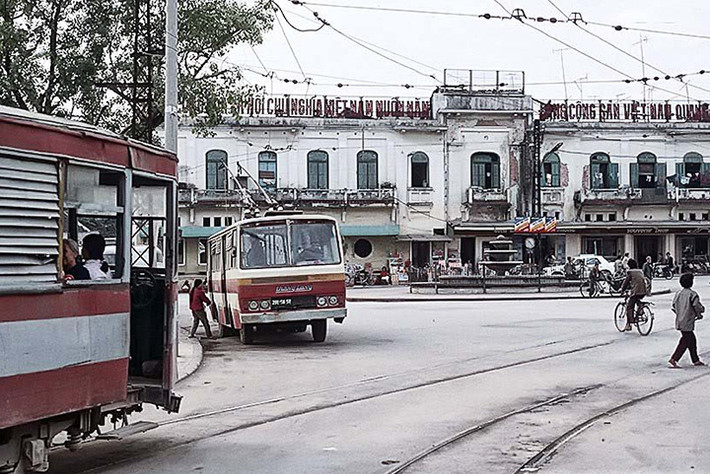 Tàu điện bánh hơi tiến vào bến tàu điện bờ hồ Hoàn Kiếm, nơi một chiếc tàu điện bánh sắt đang đỗ trên đường ray.
