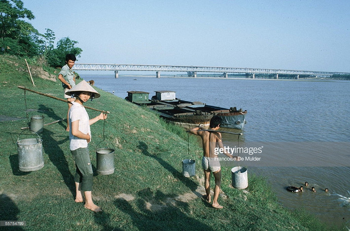 Người dân gánh nước tại bãi sông Hồng gần cầu Chương Dương. Hình ảnh nằm trong loạt ảnh Việt Nam năm 1985 của nữ phóng viên ảnh người Pháp nổi tiếng Francoise De Mulder.