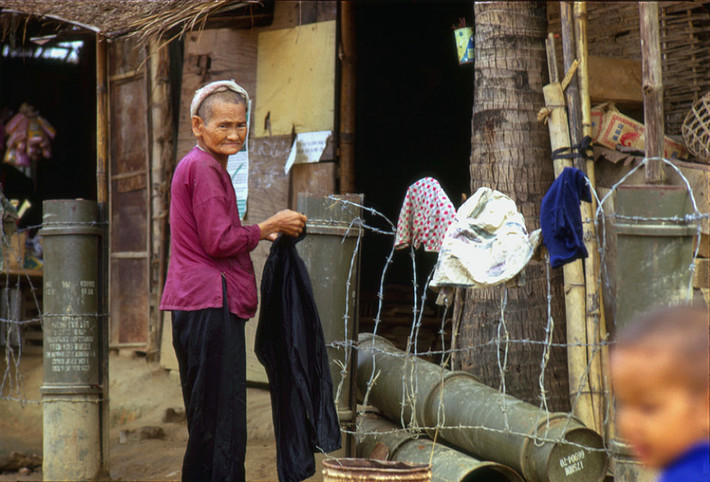 Cụ bà bên ngoài ngôi nhà có hàng rào dựng bằng vỏ bình xăng của quân đội Mỹ. Ảnh: Mark Melnick.