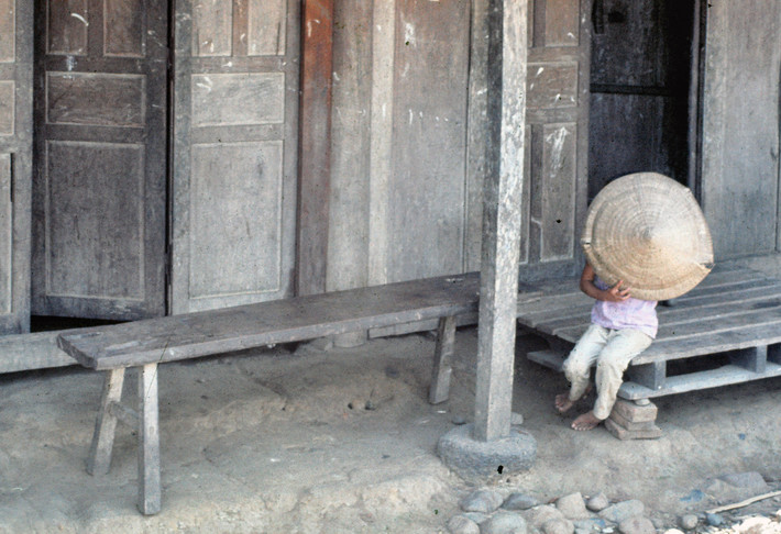 Em bé bên hiên nhà lấy nón che mặt khi được chụp hình. Ảnh: Edward Palm.