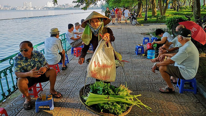Một phụ nữ gánh hàng rau cho kịp buổi chợ sớm đi qua những người đàn ông đang trầm tư bên chén chè sớm ven Hồ Tây.