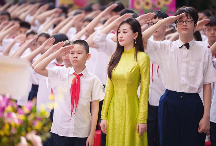 Đứng dưới sân trường, cô giáo có gương mặt thanh tú diện áo dài truyền thống màu vàng chanh nổi bật. Mái tóc dài thướt tha và lối trang điểm nhẹ nhàng càng tôn nên vẻ thanh lịch, dịu dàng.