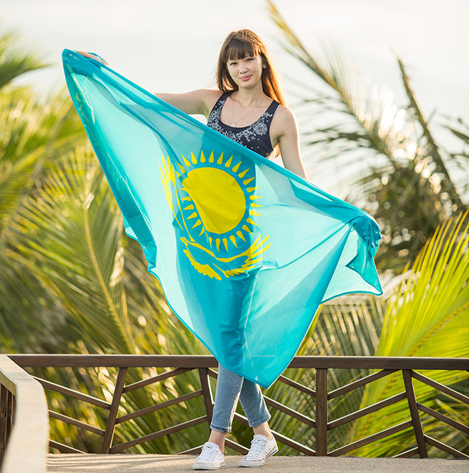 Sabina trong trang phục cá tính, giản dị trên tay cầm lá quốc kỳ Kazakhstan chụp trong khung cảnh tuyệt đẹp gần bờ biển.