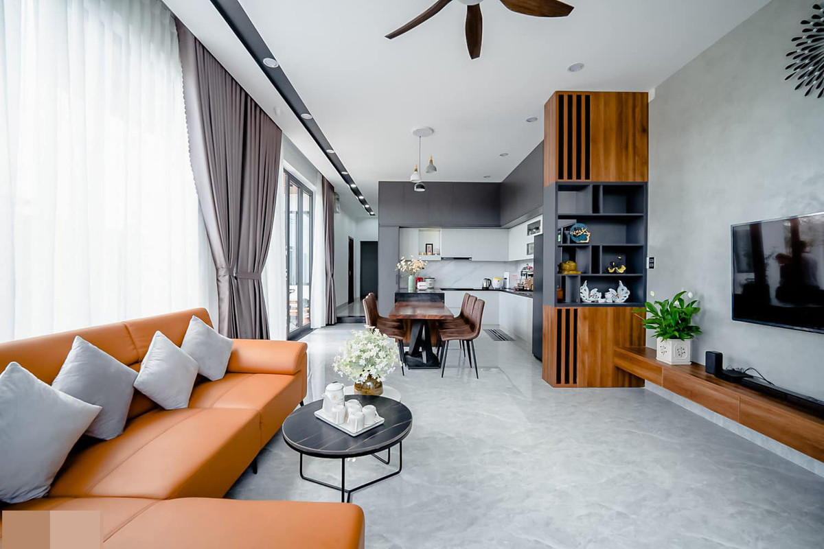 Phòng khách của ngôi nhà có nhiều khoảng sáng nhờ tường kính và cửa kính lớn. Nội thất hiện đại kiểu Hàn Quốc. Sàn lát gạch bông sáng màu dễ vệ sinh và rất mát mẻ, phù hợp thời tiết nắng nóng ở Quảng Nam. Bên cạnh phòng khách là bàn ăn và bếp nấu.