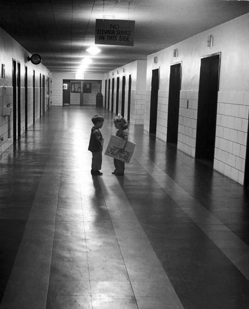 Trên hành lang trường học, hai đứa trẻ 3 tuổi dừng lại nói chuyện. Bé gái mang theo một tấm áp phích mời các bạn cùng trường đến dự triển lãm do lớp tổ chức.