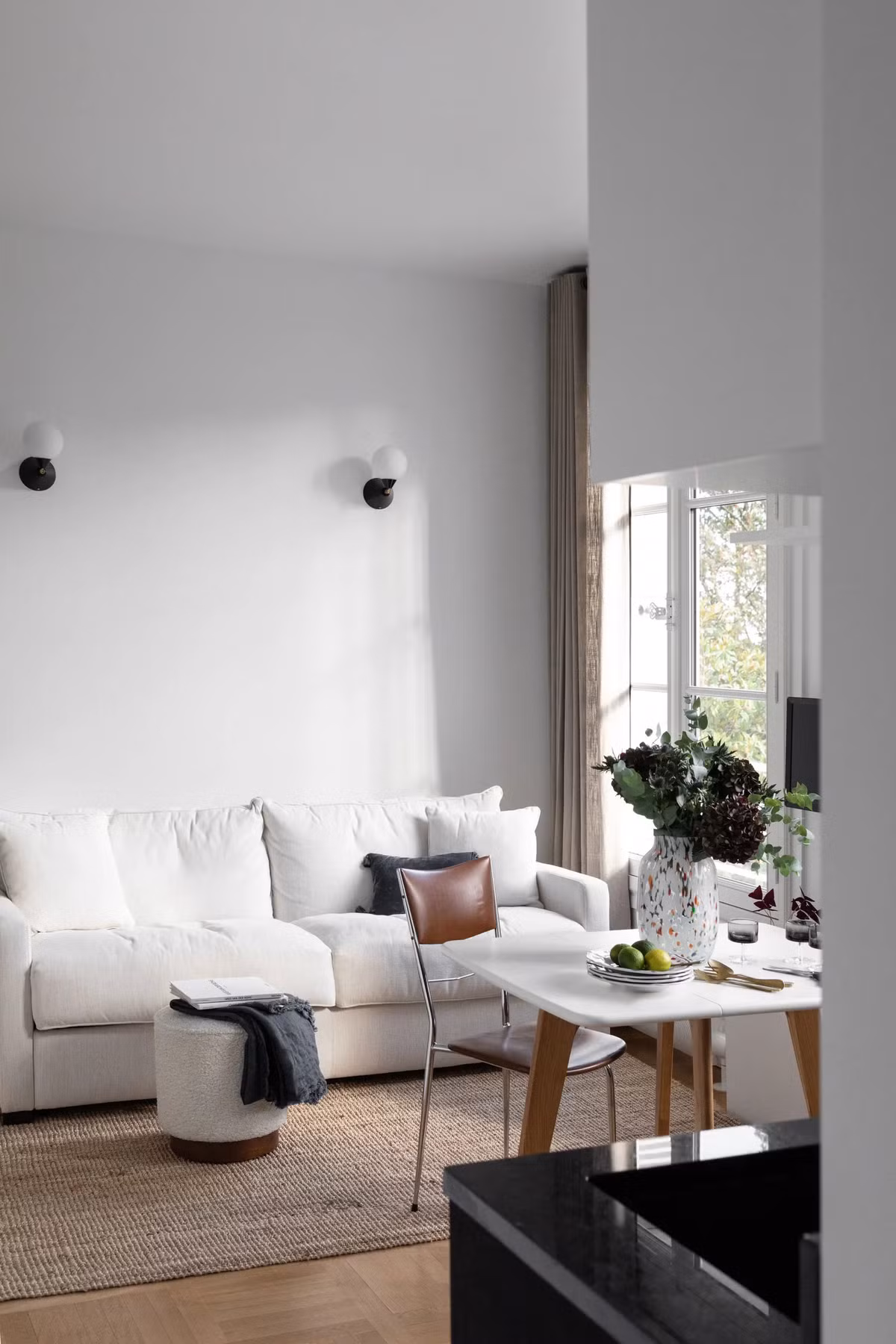 Bàn ăn vuông nhỏ xinh gần như được đặt tại giữa căn hộ nhưng lệch về phía cửa sổ. Kiểu dáng bàn ăn rất đơn giản, đơn sắc trắng. Cũng bởi thế mà chỉ cần đặt một bình hoa tươi trên đó thôi cũng đủ để toát lên vẻ đẹp sinh động, đầy sức sống. Sở hữu diện tích 25m² nên gần như mọi khu vực sinh hoạt đều diễn ra tại cùng một căn phòng. Có thể xem khu vực phòng khách được gói gọn trong tấm thảm trải sàn. 