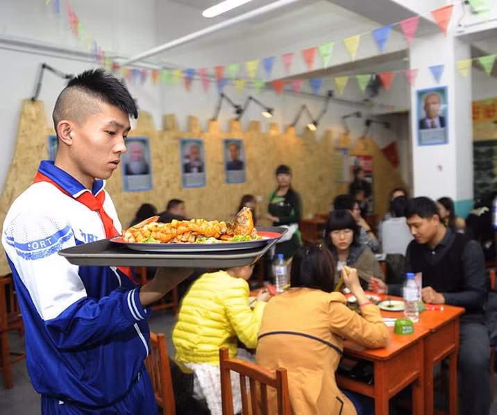 Bàn ghế được sắp xếp theo kiểu lớp học, những người phục vụ tại nhà hàng đều mặc đồng phục học sinh và đeo khăn quàng đỏ.