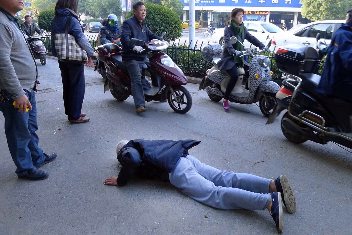 Cụ già bị ngã nằm sấp trên đường, nhiều người trông thấy nhưng rồi lại bỏ đi, chỉ còn một vài người dừng lại theo dõi.