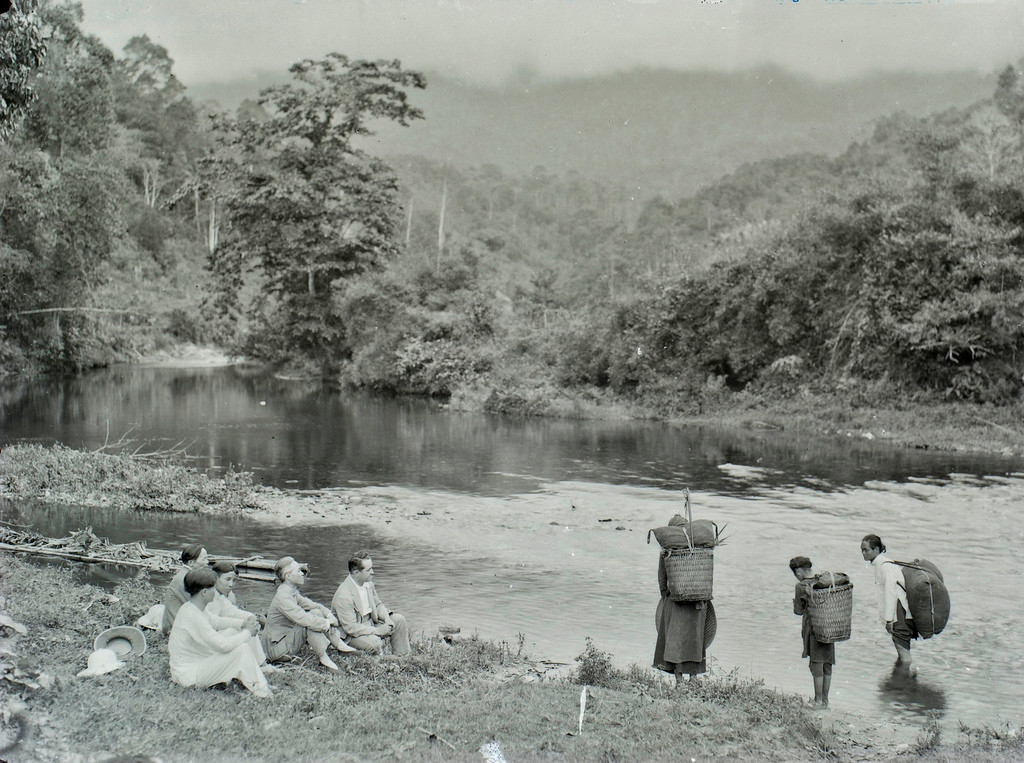 Người đàn ông Pháp cùng đoàn tùy tùng người Việt nghỉ chân bên bờ sông, gần đó là một nhóm người dân tộc thiểu số.