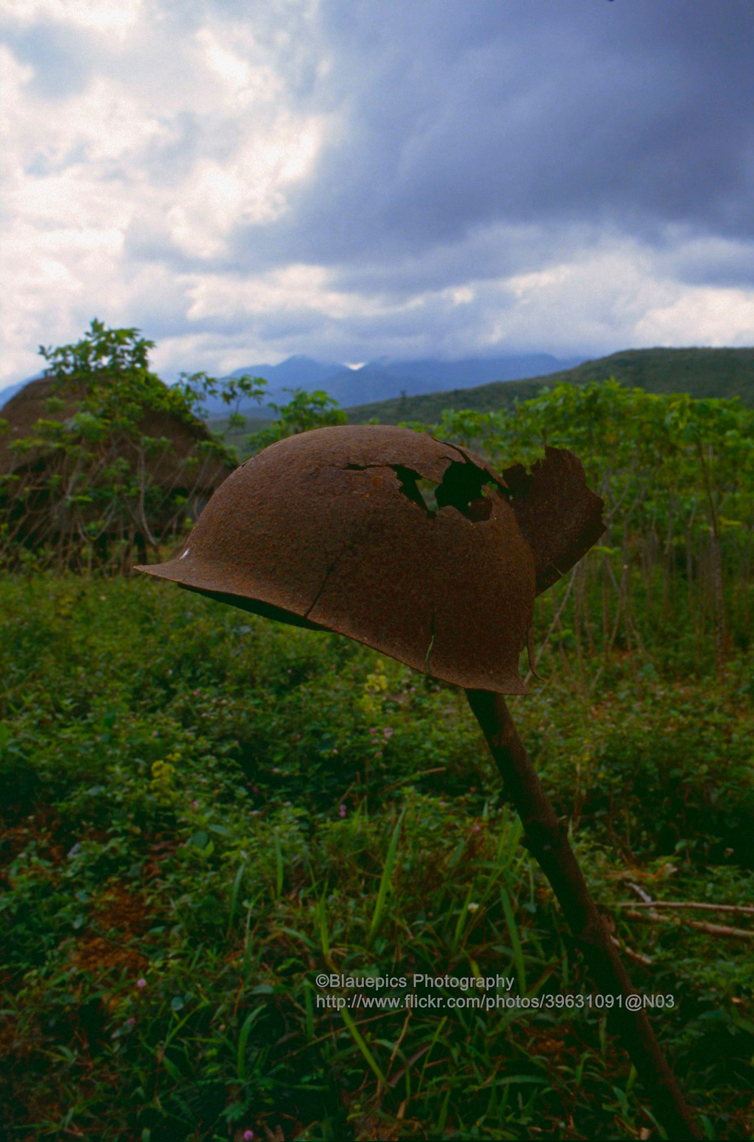 Một chiếc mũ sắt không còn nguyên vẹn được cắm trên cây cọc giữa bãi đất trống.