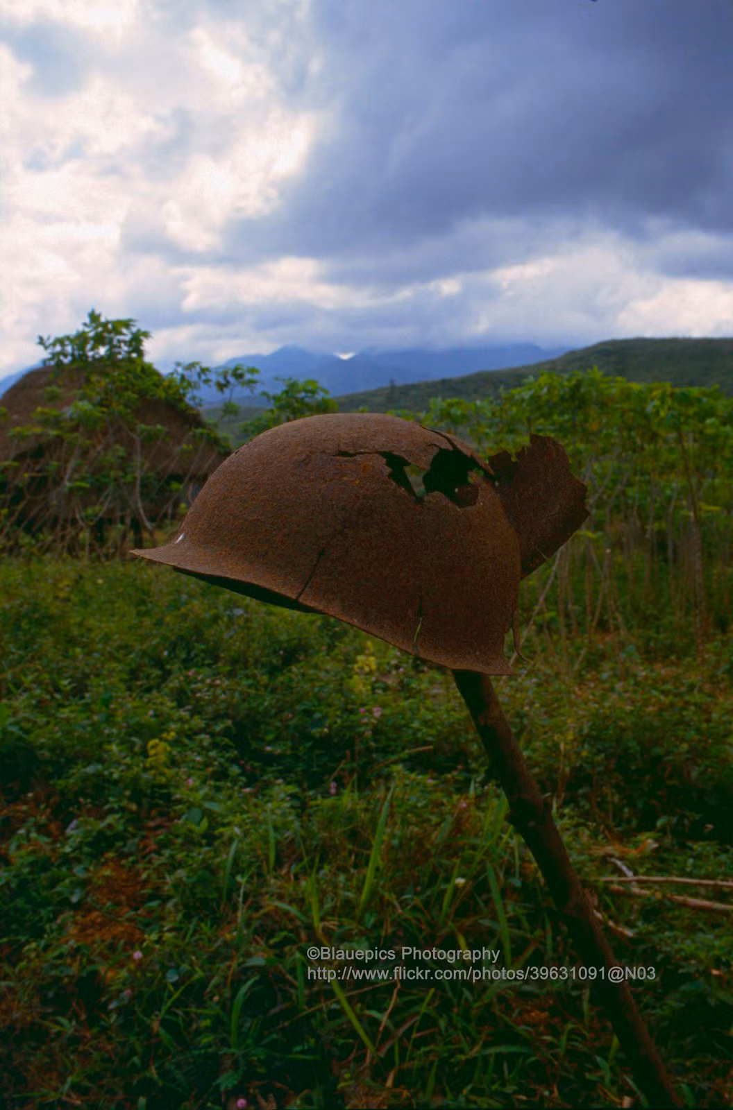Một chiếc mũ sắt không còn nguyên vẹn được cắm trên cây cọc giữa bãi đất trống.
