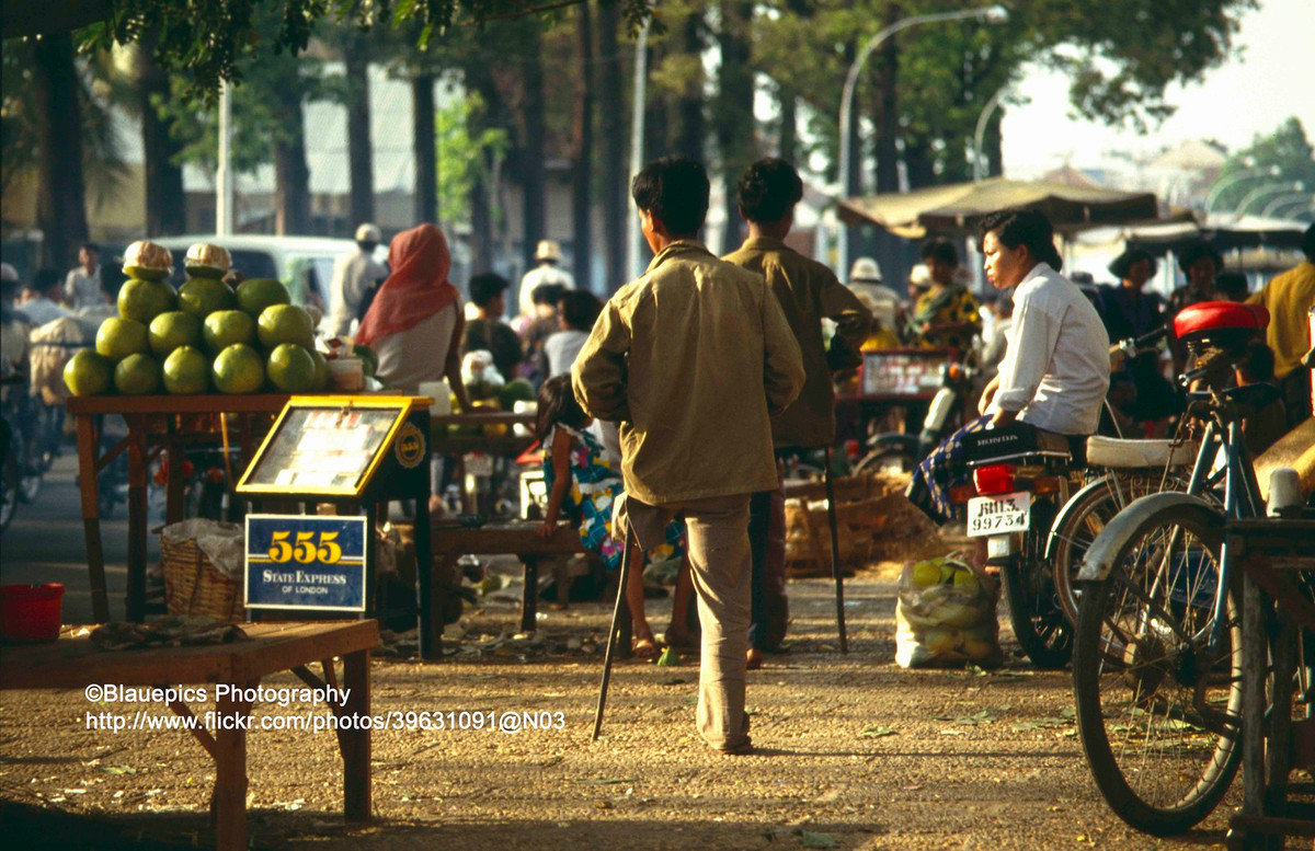 Khu chợ cóc họp trên vỉa hè thành phố Phnom Penh. Hai nam giới chống nạng đều bị mất một chân, là nạn nhân của bom mìn sót lại sau chiến tranh.