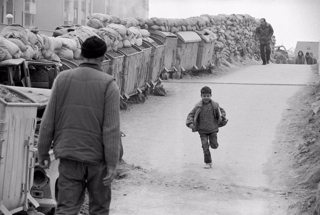 Xe rác và bao cát được dựng trên cầu Tử Thần ở quận Dobrinja, Sarajevo để chống đạn bắn tỉa từ xa, 1993. Người lớn đi qua đây thường cúi đầu thấp để bảo đảm an toàn, trong khi trẻ em thì không cần làm vậy. Ảnh: A. Abbas/ magnum Photos.