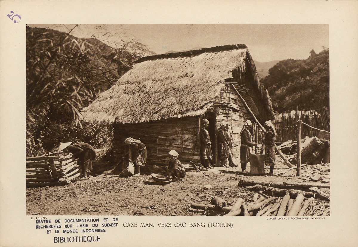 Nhà làm bằng tre nứa của người dân tộc Mán, gần Cao Bằng, miền Bắc Việt Nam. Hai người phụ nữ bên phải đang vo gạo trong chiếc cối gỗ cao. Ở giữa, một người phụ nữ đang cúi mình kiểm tra hạt ngũ cốc. Bên trái, một người đàn ông hái rau trong khu vườn nhỏ có hàng rào bao quanh.