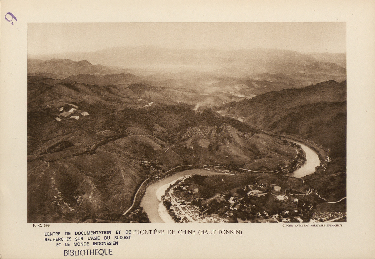 Sông Nậm Thi. Bên bờ sông là khu dân cư ngoại vi Lào Cai với nhiều nhà mái ngói dọc con đường lớn và dinh thự kiểu châu Âu trên các quả đồi.