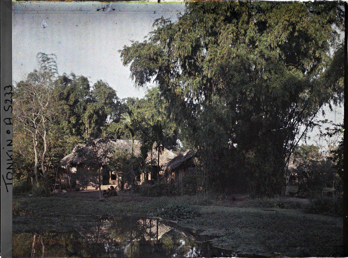 Ao nước, bụi tre và những ngôi nhà lá. Người phụ nữ và hai đứa ngồi bên bờ ao.