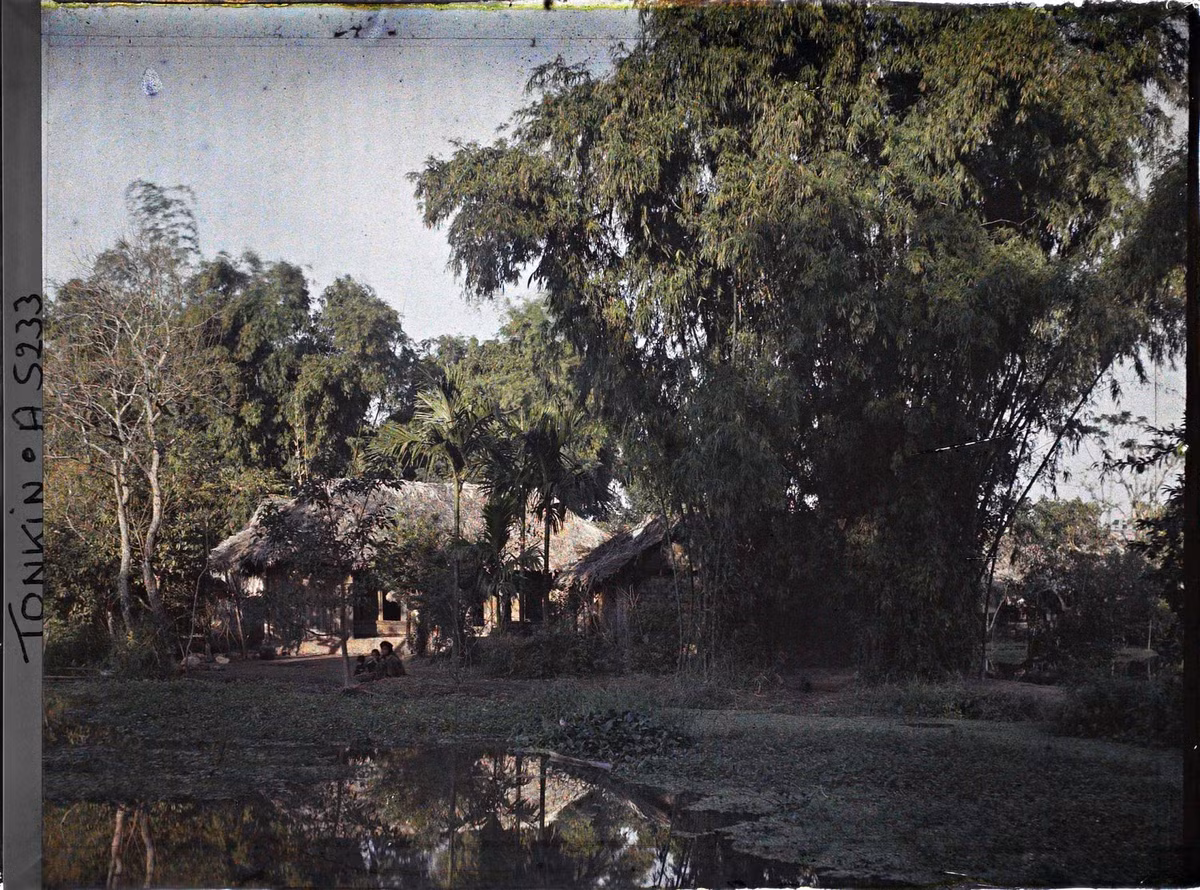 Ao nước, bụi tre và những ngôi nhà lá. Người phụ nữ và hai đứa ngồi bên bờ ao.